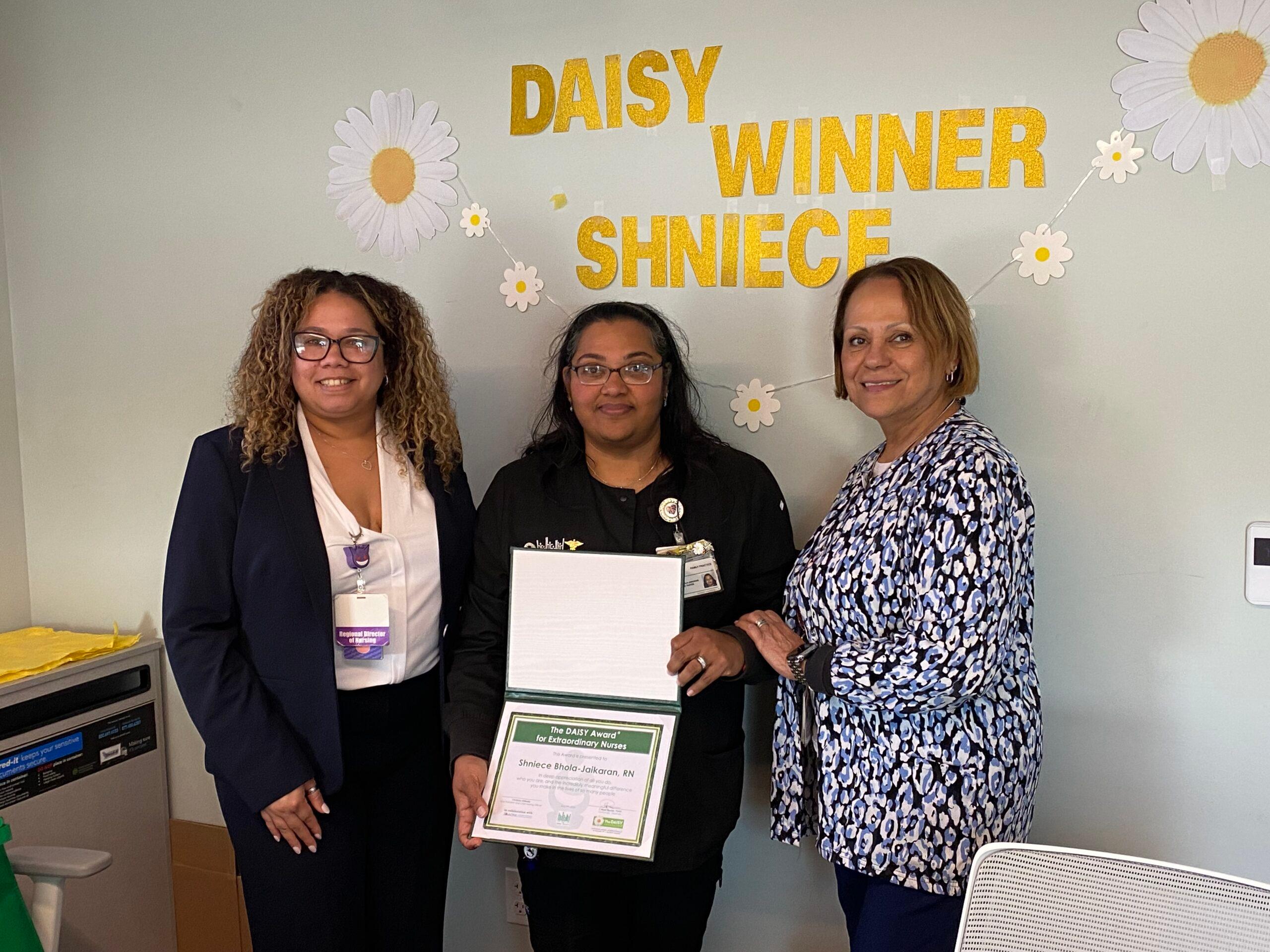 Three nurses standing against a gray wall with yellow text reading "DAISY Winner Shniece." The middle figure holds a certificate.